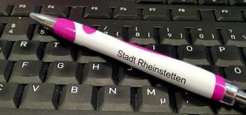 Einn weiß-rosa Kugelschreiber mit der Aufschrift "Stadt Rheinstetten " liegt quer auf einer schwarzen Tastatur 